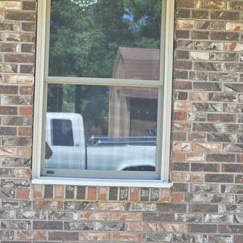 Winchoice - A rectangular window with a white frame is set into a brick wall exterior. The window reflects a white truck parked outside and trees with green foliage, along with a small building with a sloped roof. The image captures the outdoor scene through the window, with the brick wall providing a textured background. - Windows Installation Near Me - Mountain Pine, Arkansas
