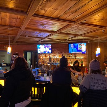 Fire Tower - a group of people sitting at a bar - The image shows the interior of a bar with a wooden paneled ceiling and a brick wall behind the bar counter. Several patrons are seated around the bar, some wearing hats and jackets, engaged in conversation or watching the TV screens. Two television screens are mounted on the brick wall, displaying different programs. The bar area has bottles and taps visible, and warm lighting illuminates the space. - New American Near Me - Stratton, Vermont