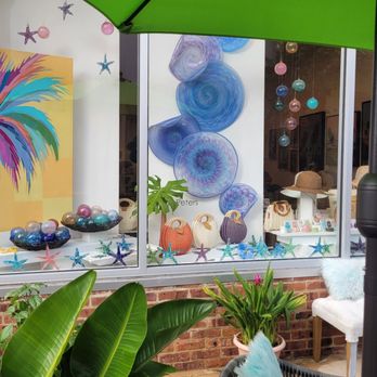 Plum Gallery - The photo captures a storefront window display with abstract blue and purple art pieces, decorative glass spheres, and woven bags arranged on the window ledge. In the foreground, there are lush green plants, a wicker chair with a blue furry pillow, and outdoor furniture. A large green umbrella partially covers the scene, and the display is set against a brick wall, indicating an outdoor seating area adjacent to the shop. - Art Galleries Near Me - Saint Augustine, Florida