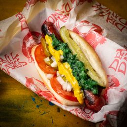 Big Chicago Dawg - A hot dog with tomato, pickle, mustard, onions, and relish in a paper wrapper, served on a wooden surface.