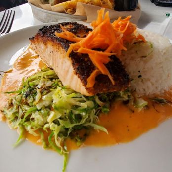 Brix Brine - Togarashi Salmon - A plate of grilled salmon with a crispy, charred skin is presented alongside a mound of white rice, shredded carrots on top, and a serving of coleslaw. The dish is accompanied by a sauce spread on the plate, and the setting includes utensils, glasses, and condiments on the table. - Seafood Near Me - Ogunquit, Maine