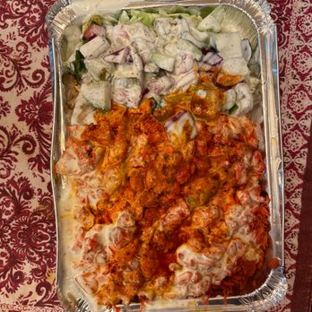 Halal Boyz - Chicken on rice - A rectangular foil tray holds a serving of chicken on rice covered with red sauce, accompanied by a side salad consisting of chopped cucumbers, onions, and a creamy dressing. The tray is placed on a patterned red and white tablecloth, suggesting a meal presentation at a restaurant or gathering. - Halal Near Me - Fresh Meadows, New York