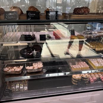 The Qahwa - Pastries - A glass display case contains various baked goods including cakes, pastries, and desserts, with some items labeled with small signs. The background shows reflections of the outside environment. - Coffee & Tea Near Me - Bridgeview, Illinois