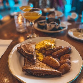 Per Diem - American Standard - A breakfast plate featuring toasted bread, scrambled eggs, sausage links, and potato wedges, with drinks and tableware in the background. - American Near Me - Lititz, Pennsylvania