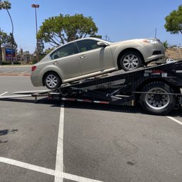 The car after getting hauled to San Diego. - A beige sedan is positioned on a flatbed tow truck in a parking lot, with trees and a shopping center visible in the background.