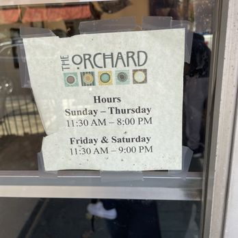 The Orchard - A printed sign taped to a glass door provides The Orchard's business hours, indicating it is open Sunday through Thursday from 11:30 AM to 8:00 PM, and Friday and Saturday from 11:30 AM to 9:00 PM. The sign features the business name and a decorative row of colored circles. The background shows reflections of the street and a person taking the photo, with some outdoor elements visible through the glass. - Seafood Near Me - Frederick, Maryland