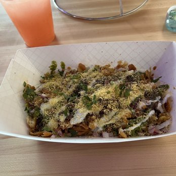 Moti Cafe - A rectangular paper tray contains loaded fries topped with shredded cheese, chopped herbs, and drizzled sauces. The tray is on a light wooden table, with a pink-colored beverage in a plastic cup and a small water bottle in the background. The setting appears to be a casual dining environment, possibly at a restaurant or food stand. - Indian Near Me - Glenview, Illinois