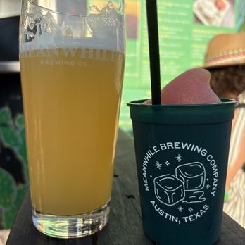 Meanwhile Brewing - A tall, clear glass of beer with foam residue at the top and a cup of pink sorbet with a black straw are positioned on a dark wooden table. The glass has the Meanwhile Brewing Co. logo, and the cup features the brewery's logo with ice cubes and stars. The background includes a green menu board and a person wearing a hat, indicating an outdoor seating area at the brewery in Austin, Texas. - Breweries Near Me - Austin, Texas