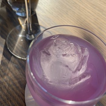 Anjou - She Wolf - A pink-colored cocktail with ice cubes is placed on a wooden table, accompanied by a glass of red wine and a glass of water in the background. - Cocktail Bars Near Me - Cincinnati, Ohio