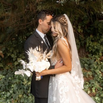 Light and Well - A bride and groom are standing close together outdoors, touching foreheads, with their eyes closed. The bride is dressed in a white lace wedding gown with a veil, and she holds a bouquet of white flowers and dried grasses. The groom is wearing a black suit with a white shirt and black tie. They are positioned under a large tree with green foliage, and the background includes dense greenery and ivy. The scene captures an intimate moment during a wedding or engagement photoshoot. - Floral Designers Near Me