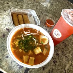 The image shows a bowl of ramen with a red broth, topped with chopped green onions, bean sprouts, tofu cubes, and half a boiled egg. Next to the ramen, there are three spring rolls in a foil container and a small cup of dipping sauce. A large pink bubble tea drink with a sealed lid and a straw is also present on the granite counter. The setting appears to be a casual dining or takeout environment.