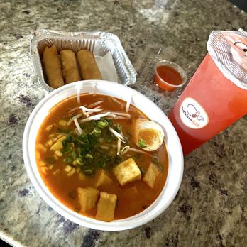 Bubblebee X Ramen - The image shows a bowl of ramen with a red broth, topped with chopped green onions, bean sprouts, tofu cubes, and half a boiled egg. Next to the ramen, there are three spring rolls in a foil container and a small cup of dipping sauce. A large pink bubble tea drink with a sealed lid and a straw is also present on the granite counter. The setting appears to be a casual dining or takeout environment. - Ramen Near Me - Ashburn, Virginia