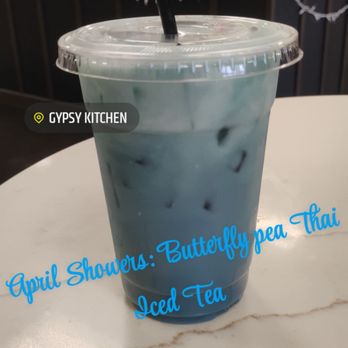 Gypsy Kitchen - Butterfly pea Thai Iced Tea - A clear plastic cup filled with Butterfly Pea Thai Iced Tea, containing ice cubes and a black straw, placed on a white table at Gypsy Kitchen. - Coffee & Tea Near Me - Lockport, New York