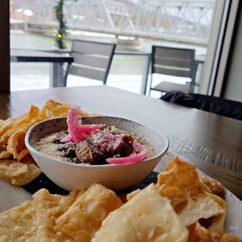Dented Keg - Brisket Queso - A bowl of brisket queso with pink pickled onions and shredded cheese, surrounded by fried chips, placed on a table by a window with river and bridge views. - Brewpubs Near Me - Clearfield, Pennsylvania