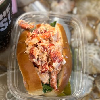 Panera Bread - Lobster roll featuring chunks of lobster meat in a toasted bun, garnished with greens, and served in a clear plastic container. The background shows a blurred surface with other food items and containers, indicating a casual or takeout setting. - Sandwiches Near Me - Needham, Massachusetts