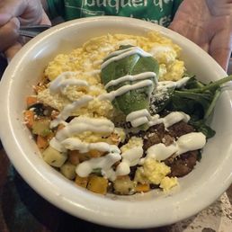 SOUTHWEST BOWL - A white ceramic bowl holds scrambled eggs, diced vegetables, leafy greens, and a drizzle of white sauce. The dish appears to be a southwestern or New Mexican cuisine, served on a dark wooden table. A person in a green shirt is holding a spoon, indicating the meal is ready to eat. The setting suggests a casual dining environment, possibly at a restaurant specializing in regional dishes.