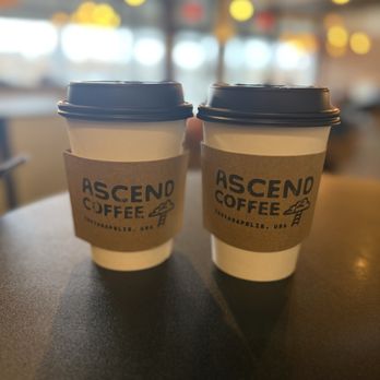 Ascend Coffee - Brown Sugar Cinnamon Latte + Brown Butter Almond Latte - Two takeaway coffee cups with Ascend Coffee branding are placed on a table inside a coffee shop, with a blurred background of the interior and lighting. - Coffee & Tea Near Me - Indianapolis, Indiana