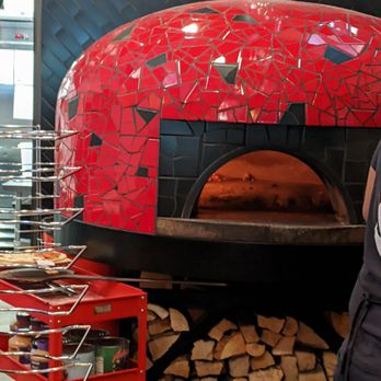 Hawt Pizza Co. - A red mosaic pizza oven with a rounded top and a black opening is depicted, with a wood storage area filled with logs underneath. The oven is situated indoors, with a dark tiled wall behind it and a metal chimney pipe extending from the top. Part of a pizza display and a person’s arm are visible in the foreground, indicating a restaurant setting. - Pizza Near Me - Albuquerque, New Mexico