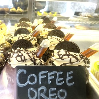 Bearded Baking - Boutique cupcakes - Display case of cupcakes topped with Oreo cookies, white frosting, chocolate drizzle, and small syringes filled with coffee, labeled 'Coffee Oreo.' - Bakeries Near Me - Manchester, New Hampshire