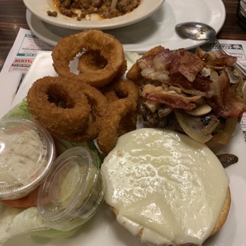 Alps Diner - A plate contains several golden-brown onion rings, a cheeseburger topped with melted cheese and crispy bacon, and a side salad with lettuce, tomato, and dressing in a small container. In the background, there is a bowl of chili with a tortilla chip on top, placed on a tray with a spoon and paper placemat. The setting appears to be a casual dining restaurant. - Diners Near Me - Wayne, New Jersey