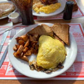 Denny's - Philly Cheesesteak Omelette - A breakfast plate includes a folded omelette, two slices of toasted bread, and a serving of breakfast potatoes. The plate is on a table with a red and white paper placemat, alongside a fork and knife. In the background, there is a glass of soda, scrambled eggs, bacon, and additional breakfast items. The setting appears to be a casual dining restaurant. - American Near Me - Lavale, Maryland