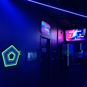 Activate Games - The image shows the interior of an arcade or gaming center with dark walls illuminated by neon blue and green lights. There are digital screens displaying game-related graphics and instructions, including one that says 'MORE GAMES' and 'CONTROL.' A group of people is gathered near the entrance or a gaming area, with some individuals taking photos or talking. The environment has a modern, high-tech aesthetic suitable for entertainment and gaming activities. - Arcades Near Me - East Rutherford, New Jersey