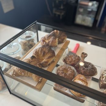 Mozz - A variety of baked pastries, such as croissants and muffins, are arranged on wooden trays within a glass display case. The display is set on a white countertop, with a blurred background of a bakery or café interior, including kitchen equipment and shelving. - Cheese Shops Near Me - Newport, Rhode Island