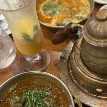 Curry Masala - Spring - Rogan Josh and Butter Chicken - The image shows a table with Indian food and beverages, including a bowl of curry garnished with herbs, a plate of butter chicken, and a cocktail with lime and mint. The dishes are served in metal and ceramic bowls, with a traditional metal teapot or container nearby. The setting appears to be a restaurant with a wooden table surface, and the food presentation suggests a casual dining experience. - Indian Near Me - Spring, Texas
