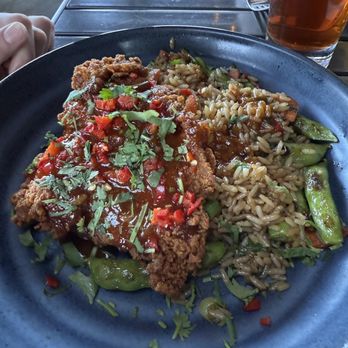 Kitchen Social - Chicken katsu - A black plate holds a serving of fried chicken covered with sauce and garnished with chopped herbs and red peppers. Next to the chicken is a portion of fried rice mixed with vegetables such as snap peas and peppers. The meal is set on a metal table with a fork, knife, and a glass of iced tea nearby, suggesting a casual dining setting. - Cocktail Bars Near Me - Columbus, Ohio