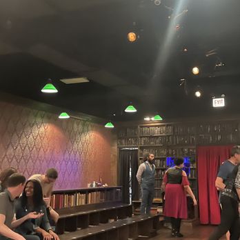 Drunk Shakespeare - Chicago - a group of people in a theatre - A group of people are seated and standing in a room with dark walls, bookshelves, and green ceiling lights, likely in a theater or performance space. - Performing Arts Near Me - Chicago, Illinois