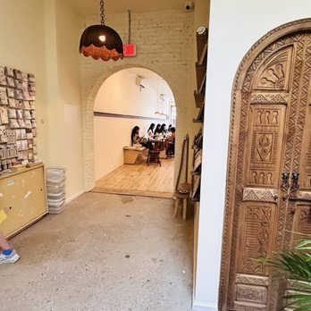 Loaf on Paper - The image shows the interior of a cafe with a carved wooden door on the right side, and a wall on the left decorated with numerous stickers or small items. There are two women near the wall, one looking at her phone and carrying a striped bag. An arched doorway leads to a seating area with several people sitting at tables. The floor is a mix of concrete and wood, and there is a small plant in the foreground. - Cards & Stationery Near Me - Brooklyn, New York