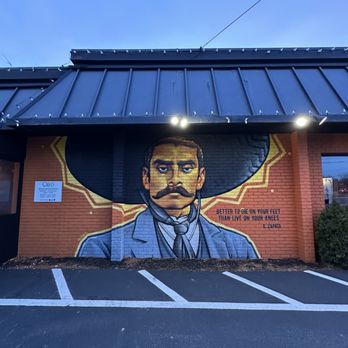 Cielo Mexican Restaurant - A mural on a brick exterior wall features a man with a large sombrero, dark hair, and a prominent mustache, dressed in a suit with a tie. The mural includes a quote attributed to Zapata: "Better to die on your feet than live on your knees." The wall is part of a building with a metal roof, and there are parking spaces in front of it. Two bright lights illuminate the mural from above, and a small sign is visible on the left side of the wall. - Mexican Near Me - Braintree, Massachusetts