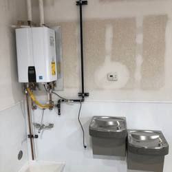 Alpine Services - The image shows a utility area with a white tankless water heater mounted on the wall. Below it is a utility sink, and to the...