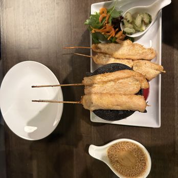 Dillon Thai restaurant - Chicken Satay special appetizer - The image shows a rectangular white plate with skewered fish and shrimp, accompanied by a small salad with mixed greens and shredded carrots, and cucumber slices in a white spoon. A small bowl of dipping sauce is placed nearby. The setting appears to be a restaurant with a dark wooden table. - Thai Near Me - Dillon, Colorado