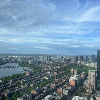 View Boston - A high-angle view of Boston's skyline, featuring a river with bridges, numerous buildings, and green spaces, under a partly cloudy sky. - Observatories Near Me - Boston, Massachusetts