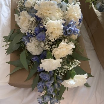 Locker's Flowers - Day of (this one was pretty just not what was originally made) - A cascading floral arrangement features white carnations, blue delphiniums, and small white filler flowers, accented with green leaves. The bouquet is wrapped in brown paper and placed on a white surface, with additional floral arrangements visible in the background. - Florists Near Me - McHenry, Illinois