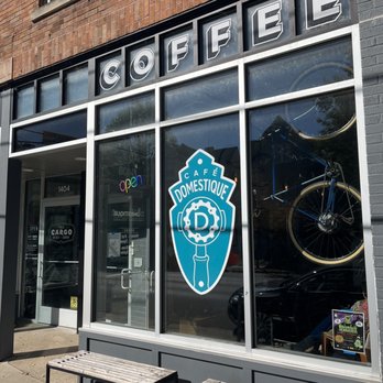 Café Domestique - the front of a coffee shop - The image shows the storefront of Café Domestique, a bicycle-themed cafe. The facade includes large glass windows with a bicycle mounted inside, visible through the glass. The cafe's logo, featuring a gear and a bicycle chainring, is displayed on the window. Above the entrance, the word 'COFFEE' is spelled out with individual letters in a modern font. Outside, there are two benches on the sidewalk, and the building's exterior is made of brick and painted gray. The entrance door has a neon 'open' sign and various notices posted on it. - Coffee & Tea Near Me - Madison, Wisconsin