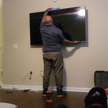 Cool Geekz - A man with tattoos is using a level tool to install or adjust a flat-screen TV on a beige wall in a living room. - Home Theatre Installation Near Me