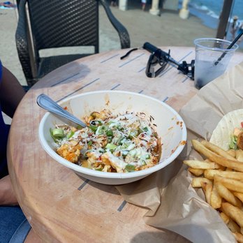 SurfSide - Bang Bang Cauliflower - A bowl of Bang Bang Cauliflower with sauce, chopped green onions, and shredded cheese is placed on a wooden table at an outdoor beachside dining area. Next to it, there is a paper-lined basket of French fries and a plastic cup with a beverage and a straw. The background shows a sandy beach, ocean, and a pier structure, indicating a seaside location. - Bars Near Me - Salisbury, Massachusetts