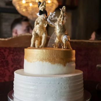 The Evercake - A wedding cake with two gold elephants as decorative toppers is displayed on a dark surface. The elephants are detailed with gold accents and are positioned facing each other. The cake has a smooth white frosting with a gold-painted top tier. In the background, a chandelier with warm lighting hangs from the ceiling, and blurred figures and furniture are visible, indicating an indoor celebration setting. - Custom Cakes Near Me