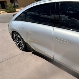 The image depicts a silver car parked on a concrete driveway, with visible scratches and dents on the lower part of the driver's side door and side panel. The car has tinted windows and stylish alloy wheels. The background includes a residential area with a gravel pathway, some plants, and a building with windows. The sky is partly cloudy, reflected on the car's windows.