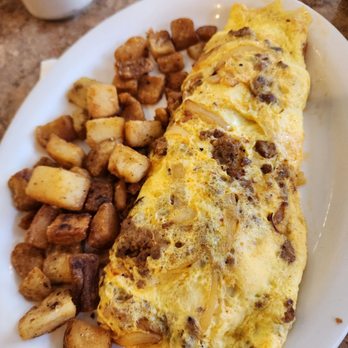 The Pantry - Soupy and fried onion omelet - The image shows a breakfast dish with a large omelet containing ground beef and onions, served on a white oval plate. Next to the omelet are fried breakfast potatoes, cut into small cubes and seasoned. The plate is placed on a brown, textured table surface, with a white mug visible in the background. - Diners Near Me - Westerly, Rhode Island