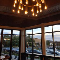 Ambiance - The image shows the interior of a cafe or restaurant with large floor-to-ceiling windows allowing natural light and views of parked cars and trees outside. A modern chandelier with multiple exposed filament bulbs hangs from the ceiling, providing warm lighting. There are bar-height seats along the window ledge and a small round table with chairs visible in the corner. The space has a contemporary design with dark flooring and ceiling accents.