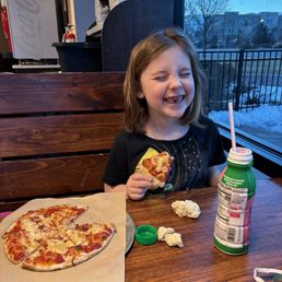 A young girl with shoulder-length brown hair is sitting at a wooden table in a restaurant, smiling with her eyes closed and holding a slice of pizza. On the table, there are two pieces of crumpled napkins, a green bottle with a straw, and a pizza on a tray with a few slices remaining. The background shows a large window with a view of trees and buildings outside, and part of the restaurant interior including a Coca-Cola machine and a fire extinguisher.