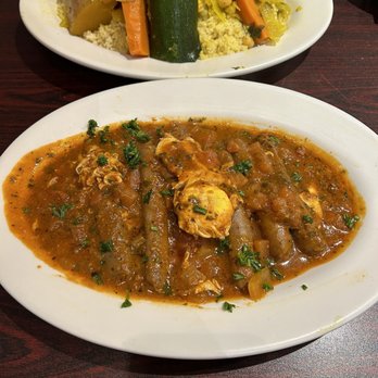 Fettoosh - Merguez Tagine - The image features two Moroccan dishes served on white plates. The top dish contains couscous topped with cooked vegetables including zucchini, carrots, and chickpeas. The bottom dish is a stew with a tomato-based sauce, vegetables, and a poached egg, garnished with chopped parsley. The dishes are placed on a dark wooden table, with a small bowl and a napkin visible in the background. - Moroccan Near Me - Arlington, Virginia