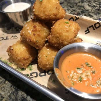 Topgolf - Mac N Cheese Bite mound of tastiness with two dipping sauces! - Fried cheese balls are stacked on a metal tray with a side of orange dipping sauce and a small bowl of white sauce, served on a restaurant table. - New American Near Me - Albuquerque, New Mexico