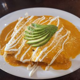 Enchiladas Suizas - A white oval plate holds Enchiladas Suizas, featuring rolled tortillas covered in orange sauce, garnished with sliced avocado on top. The dish is decorated with white sauce drizzles and a sprinkle of cheese, placed on a dark wooden table.