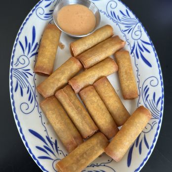 Cebu Chicago - Lumpia - Fifteen fried spring rolls are arranged on an oval ceramic plate with a blue floral pattern. The spring rolls are golden brown and cylindrical. A small metal cup of dipping sauce, likely peanut or sweet chili, is placed at the top of the plate. The plate is set on a dark surface, and the image captures the food presentation clearly. - Filipino Near Me - Chicago, Illinois