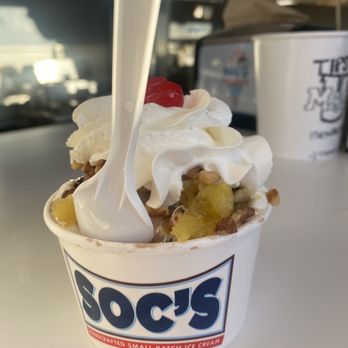 Soc's Ice Cream - Hot Fudge Sundae w/pineapple and Chocolate Hard Ice Cream - A cup of ice cream from Soc's Ice Cream features whipped cream, a cherry, and fruit toppings, with a plastic spoon inserted. - Ice Cream & Frozen Yogurt Near Me - Saugus, Massachusetts