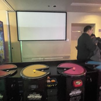 ENTERRIUM - lots of broken/out of order games - Arcade game machines with colorful controls are positioned in front of a blank screen in a dimly lit entertainment venue. - New American Near Me - Schaumburg, Illinois